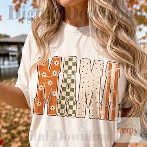 May include: A cream-colored short-sleeve t-shirt featuring the word MAMA in large block letters. The letters have a distressed texture with varied patterns: the first M has white daisies on orange, the A is green checkered, and the MA has polka dots.