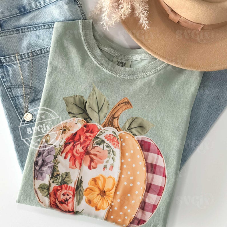 May include: A light green t-shirt featuring a patchwork pumpkin design with floral, polka dot, and plaid patterns. The shirt is paired with blue jeans, a gold necklace, and a tan hat, creating a fall-themed ensemble.