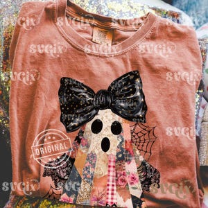 May include: A terracotta-colored t-shirt featuring a ghost design with a black bow and patchwork body. The ghost has three black eye holes and is decorated with a spiderweb. The word "ORIGINAL" is stamped on the shirt.