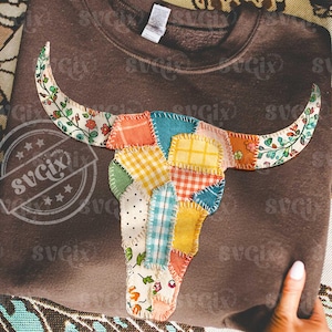 Może przedstawiać: Brązowa bluza z kapturem z patchworkowym wzorem czaszki byka. Czaszka składa się z różnych wzorów tkanin, w tym kwiatowych, w kratkę i w kropki. Rogi zdobione są kwiatową tkaniną. Projekt jest wyśrodkowany na bluzie.