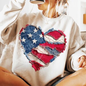 May include: A cream-coloured sweatshirt with a heart-shaped design made from denim and red fabric patches. The blue denim section features white stars. The edges of the patches are frayed, giving a rustic appearance. A casual, patriotic style.