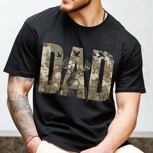 May include: Black t-shirt with a camouflage pattern featuring the word "DAD" with a deer silhouette in the middle of the word.