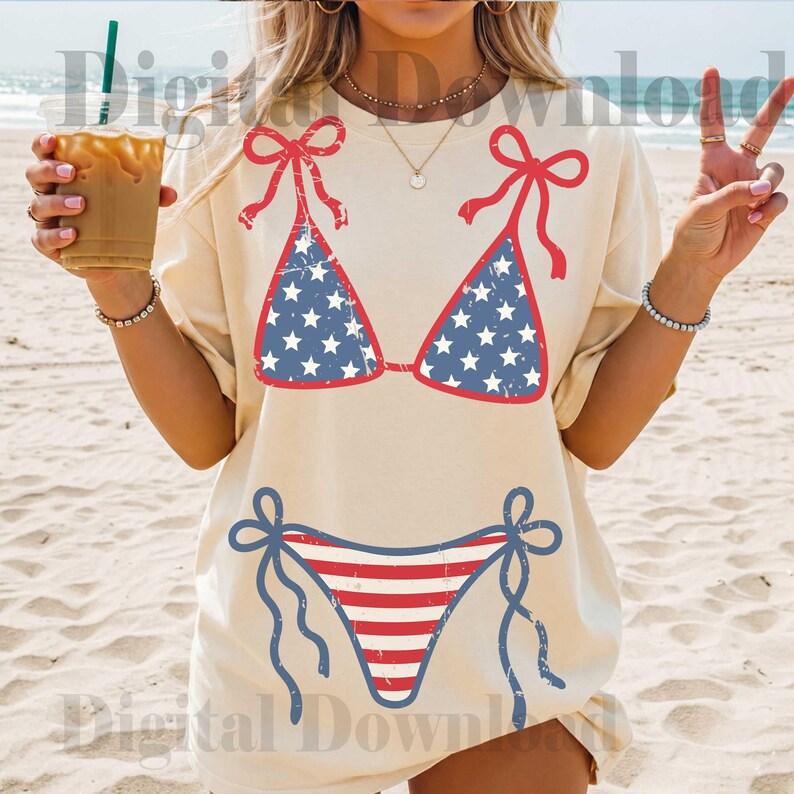 May include: Cream-colored t-shirt featuring a patriotic bikini design. The bikini top is blue with white stars, and the bottom is red and white striped. Red bows accent the top and bottom. The shirt is worn on a beach.