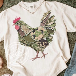 Può includere: T-shirt color crema con un'applicazione dettagliata di un gallo in tessuto mimetico. Il gallo ha cresta e bargigli rossi, con una collana d'argento allo scollo. La maglietta è di Comfort Colors.