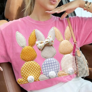 May include: Pink t-shirt featuring three appliqued bunny rabbits. The bunnies have white, yellow, and light orange polka-dotted and patterned bodies and ears. One bunny has a floral bow. The bunnies have white fluffy tails.