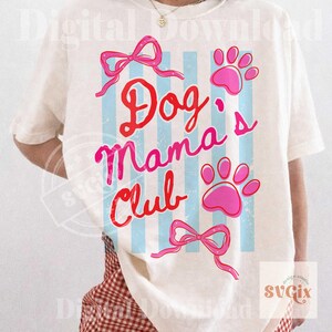 May include: Cream-colored t-shirt with the text "Dog Mama's Club" in red and pink script. The design includes pink paw prints, bows, and blue vertical stripes. A casual, pet-themed graphic tee.