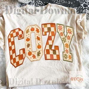 May include: A cream-colored t-shirt featuring the word COZY printed in large letters. The C is a brown and white checkerboard, the O contains orange pumpkins and leaves, the Z is a brown and white checkerboard, and the Y has orange flowers.