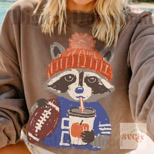 May include: A brown sweatshirt featuring a graphic of a raccoon wearing an orange knit hat, holding a brown football, and drinking from a white cup with an orange pumpkin illustration. The design has a distressed, textured appearance.