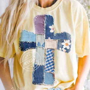 Op de afbeelding: Een lichtgeel t-shirt met een patchwork kruisontwerp. Het kruis is gemaakt van verschillende denimstoffen, waaronder blauwe bloemen- en ruitpatronen en lichtpaarse stof. Het kruis is op het shirt gestikt.