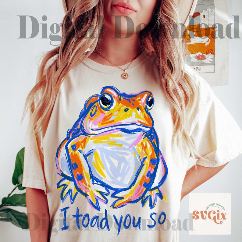 May include: Cream-colored t-shirt featuring a colorful, artistic illustration of a frog. The frog is depicted in shades of blue, orange, yellow, and pink. Below the frog, the text reads "I toad you so".