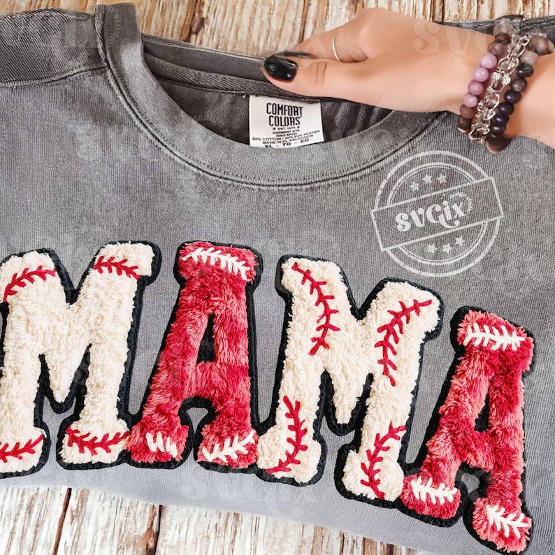 May include: Gray sweatshirt with the word "MAMA" in baseball-themed lettering. The letters are white and red, with red stitching details. The sweatshirt has a Comfort Colors tag.