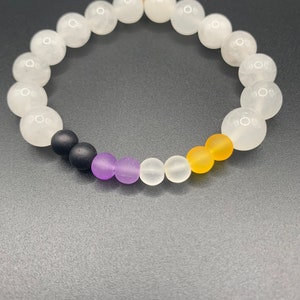 Könnte beinhalten: Ein weißes Perlenarmband mit einer schwarzen, violetten, weißen und gelben Akzentperle. Das Armband besteht aus runden Perlen und ist etwa 20 cm lang.