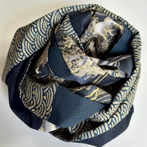 May include: A rolled scarf with a patchwork design. It features navy blue, gold, and off-white colours. The patterns include wave-like motifs, small dots, and floral designs. The scarf is made of a soft fabric. The scarf is a stylish accessory.