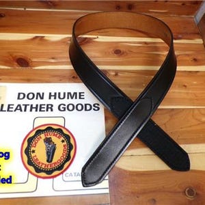 Może przedstawiać: Czarny skórzany pasek z brązowym wnętrzem leży na katalogu. Na katalogu widnieje napis "DON HUME LEATHER GOODS" z okrągłym logo. Widoczny jest również tekst "Catalog Not Included".