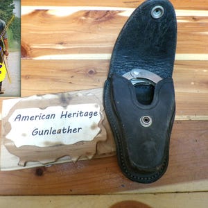 May include: Black leather tear drop cuff case with a silver handcuff inside. The case has a snap closure and is displayed on a wooden surface. A sign reads "American Heritage Gunleather".