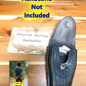 May include: Black leather Tear Drop Cuff Case with a silver handcuff inside. The case has a decorative pattern and a snap closure. The text "Handcuffs Not Included" is in yellow and blue. The text "American Heritage Gunleather" is on a wooden background.