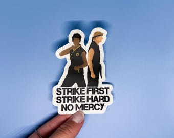 Puede incluir: Una pegatina con dos figuras en uniformes de karate negros con el texto "Strike First Strike Hard No Mercy".
