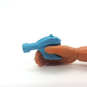 Könnte beinhalten: Eine hellblaue Spielzeugpistole aus Plastik mit einem schwarzen Lauf und einem silbernen Detail oben. Die Pistole wird in einer braunen Plastikhand gehalten.