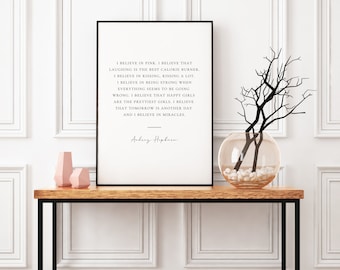 Pode incluir: Uma impressão emoldurada com uma borda preta exibe uma citação atribuída a Audrey Hepburn. A citação está em texto preto sobre um fundo branco. Uma mesa de madeira contém a impressão emoldurada, dois vasos geométricos rosa e um vaso de vidro com galhos.