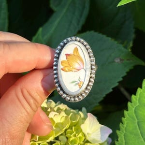 May include: An oval ring featuring a white ceramic inlay with a hand-painted yellow and orange tulip design. The center piece is set within a textured, beaded sterling silver frame that creates a distinct border around the floral illustration.