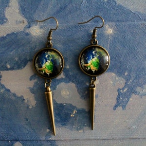May include: A pair of dangle earrings with a bronze finish. Each earring features a round glass cabochon with a colorful image of the Earth. The cabochons are attached to a long, pointed bronze pendant.