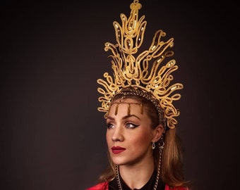 May include: A woman wearing a red and gold patterned robe with a gold crown and a gold chain necklace. She is looking to the side and has her hands clasped in her lap.
