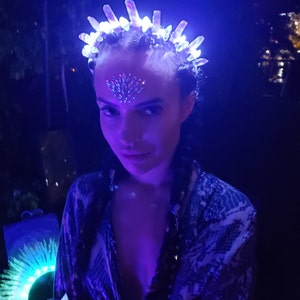 May include: A woman wearing a white and blue patterned shirt with a crystal headband that has blue lights. The headband is on top of her head and has a crown shape. She is wearing makeup with rhinestones on her forehead.