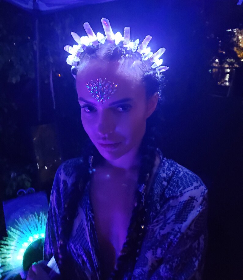 May include: A woman wearing a white and blue patterned shirt with a crystal headband that has blue lights. The headband is on top of her head and has a crown shape. She is wearing makeup with rhinestones on her forehead.