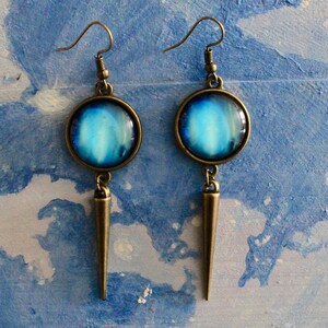 May include: A pair of dangle earrings with a blue and white galaxy design in a round glass cabochon. The earrings have a bronze finish and a long, pointed bronze dangle.