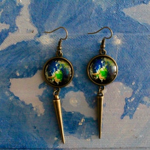 May include: A pair of dangle earrings with a bronze finish. Each earring features a round glass cabochon with a painted image of the Earth. The cabochons are attached to a long, pointed bronze pendant.