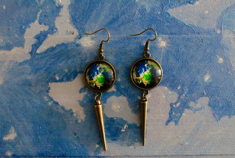May include: A pair of dangle earrings with a bronze finish. Each earring features a round glass cabochon with a painted image of the Earth. The cabochons are attached to a long, pointed bronze pendant.