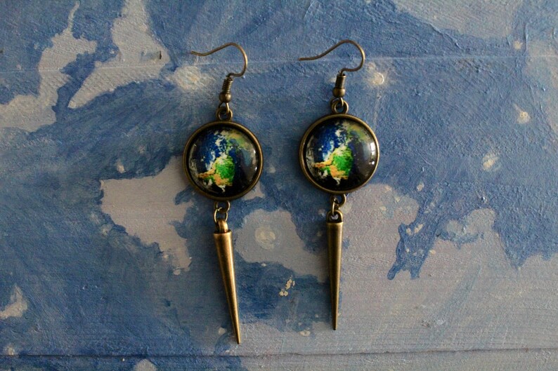 May include: A pair of dangle earrings with a bronze finish. Each earring features a round glass cabochon with a colorful image of the Earth. The cabochons are attached to a long, pointed bronze pendant.
