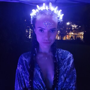 May include: A woman wearing a white crystal crown with lights, a silver nose ring, and a blue and white patterned shirt.