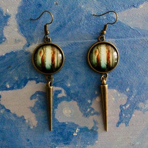 May include: A pair of long, bronze earrings with a round, glass pendant featuring a colorful abstract design. The pendants are attached to a long, pointed bronze dangle.