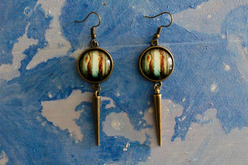 May include: A pair of long, bronze earrings with a round, glass pendant featuring a colorful abstract design. The pendants are attached to a long, pointed bronze dangle.