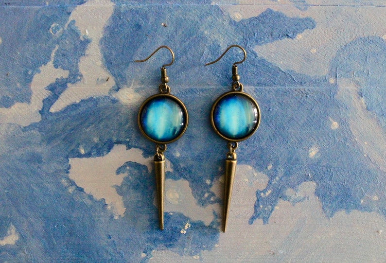 May include: A pair of dangle earrings with a blue and white galaxy design. The earrings have a bronze finish and a pointed spike hanging from the bottom.
