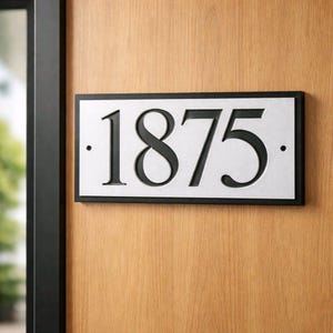 Op de afbeelding: Rechthoekig adresbord met het nummer 1875 in zwart op een zilveren achtergrond. Het bord is zwart omlijst en bevestigd op een houten deur. De cijfers zijn in een klassiek schreeflettertype.
