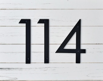 Può includere: Numeri civici neri in metallo, 114, su sfondo in legno bianco. I numeri sono in stile moderno, senza grazie, e montati con viti. I numeri presentano un design audace e minimalista.