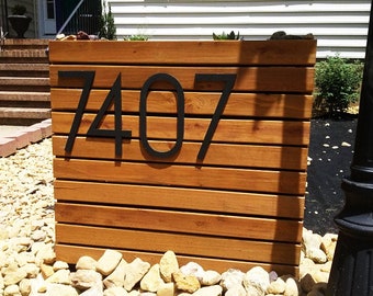 Puede incluir: Un letrero de número de casa de madera con números de metal negro. Los números son 7407. El letrero está rodeado de pequeñas piedras.