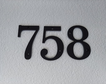 May include: Black metal house numbers 758 on a white textured wall.
