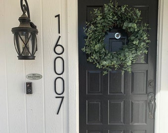 May include: Black metal house numbers 16007 are mounted on a white wall. A black door with a wreath of green leaves is to the right of the numbers. A doorbell and a sign that reads "No Soliciting" are below the numbers.