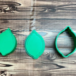 May include: Three bright green plastic cookie cutters shaped like lemons. Two of the cutters are solid pieces with embossed details on the surface, while the third cutter is an open frame designed to cut the outline of the lemon shape.