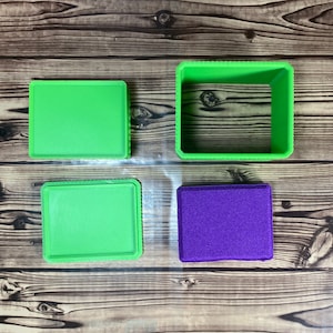May include: Four square boxes in green and purple. Two of the boxes are open and empty. The other two boxes are closed. The boxes are on a wooden surface.
