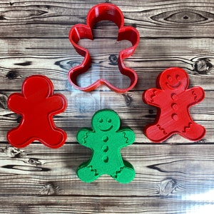 May include: Three gingerbread man cookie cutters, one red, one green, and one red with a smiling face and buttons. The cutters are on a wooden surface.