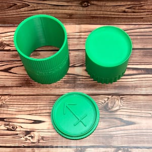May include: A bright green cylindrical container with a ribbed exterior texture, consisting of an open-ended tube, a solid cap, and a separate circular lid featuring a raised Sagittarius zodiac arrow symbol embossed on the flat top surface.