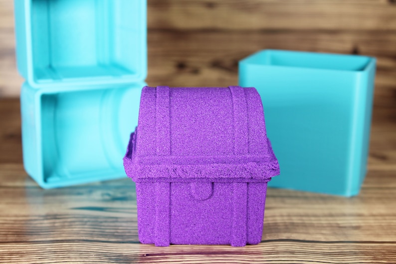 以下が含まれることがあります： 紫色の砂でできた宝箱の形をした砂のお城。宝箱は蝶番のついた蓋が付いており、木製の表面の上に置かれています。