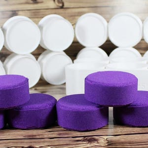 May include: A collection of vibrant purple, cylindrical bath bombs arranged in the foreground, accompanied by a white plastic multi-cavity mold tray positioned behind them. The mold tray features several empty circular compartments for shaping.