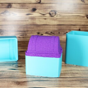 以下が含まれることがあります： ターコイズブルーのプラスチック製の容器が3つ。1つは紫色の砂のような蓋が付いた宝箱の形、もう2つは正方形の容器です。