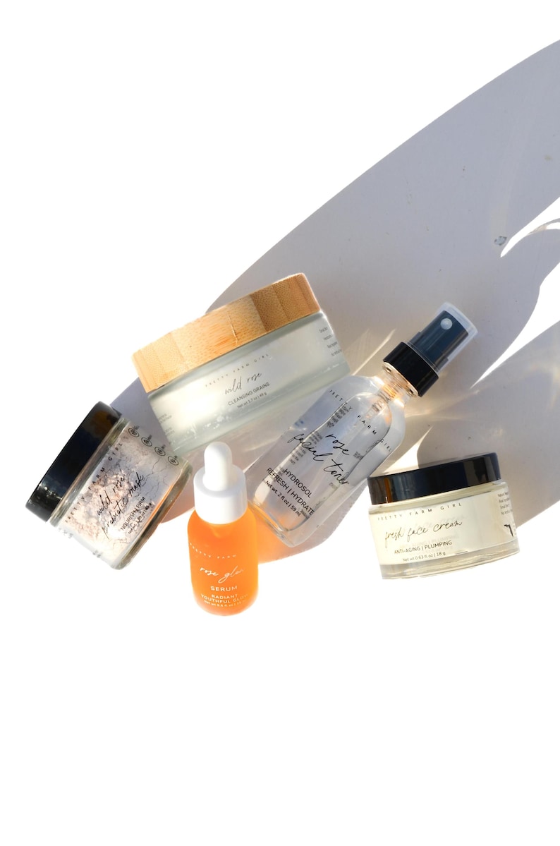 Puede incluir: Una colecci&oacute;n de cinco productos para el cuidado de la piel en frascos y botellas de vidrio con etiquetas blancas. Los productos incluyen un b&aacute;lsamo limpiador, un suero facial de rosa, un t&oacute;nico de agua de rosas, una crema fresca para la cara y una mascarilla de arcilla desintoxicante. Los productos est&aacute;n dispuestos sobre una superficie blanca.