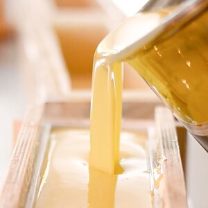 May include: A close-up of a clear glass pitcher pouring a golden yellow liquid into a wooden mold. The liquid is thick and viscous, and it is filling the mold to the top.